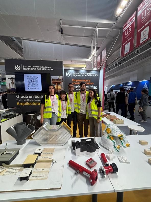 Fotografía de 5 estudiantes en el stand de Edificación. Visten con chalecos de seguridad y cascos, y están posando tras una mesa llena de utensilios propios de la profesión (maquetas, tuberías, ladrillos, etc)