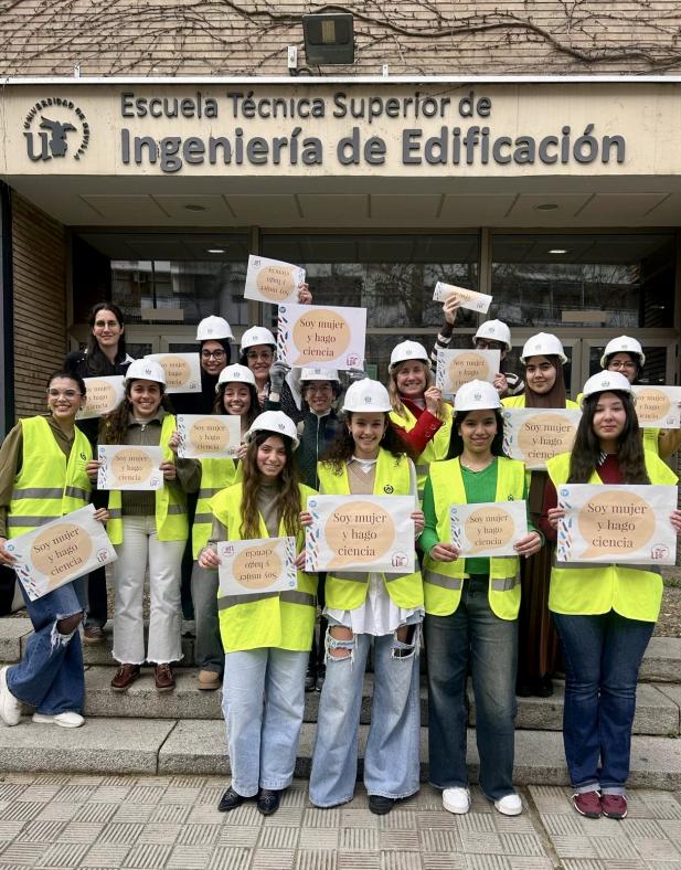 Fotografía de profesoras y alumnas de la ETSIE vistiendo casco y chaleco de seguridad mientras sostienen un cartel que dice "soy mujer y hago ciencia". Unicación de la fotografía en la puerta principal de la ETSIE.