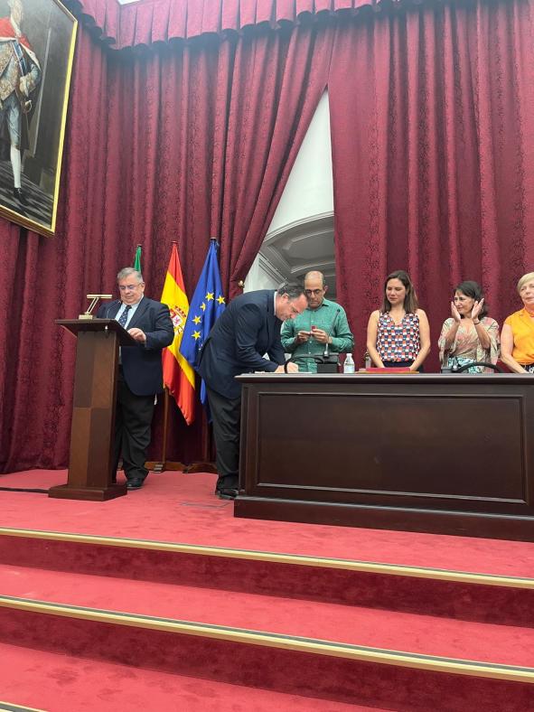 Fotografía de D. Juan Jesús Martín Del Río firmando su toma de posesión en el paraninfo de la Universidad de Sevilla. Al fondo se ven las banderas de Andalucía, España y la Unión Europea.
