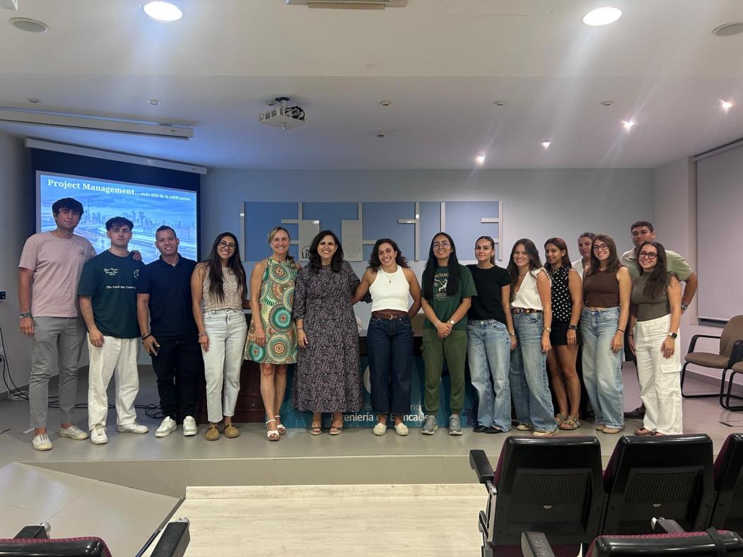 Fotografía de Macarena Parra junto a la Directora Charo Chaza y parte del alumnado asistente. De fondo las palabras ETSIE en la pared del salón de Grados.